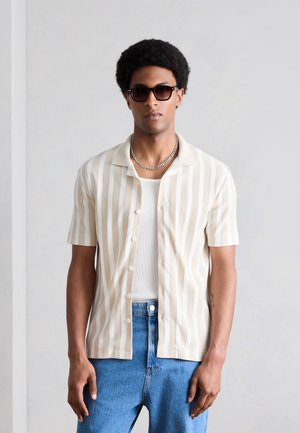 Άνδρας με γυαλιά ηλίου, πουκάμισο με κάθετες μπεζ και λευκές ρίγες με κοντά μανίκια πάνω από λευκό αμάνικο, μπλε τζιν και ασημένιο αλυσίδα λαιμού.