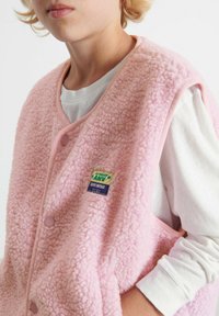 Różowa kamizelka polarowa o teksturowanej, puszystej powierzchni, z okrągłym dekoltem, zapięciem na guziki oraz kolorową metką marki z przodu.