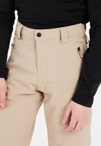 Pantaloni impermeabili beige con vita alta, tasche a toppa e cerniere sui lati. Texture liscia, tessuto robusto e chiusura con bottone nero.