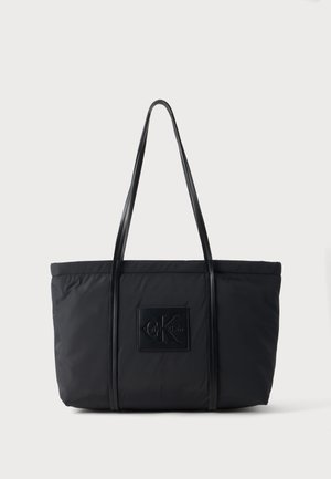 Sort Calvin Klein tote taske med lange stropper og logopatch på fronten, sat mod en ensfarvet lys baggrund.