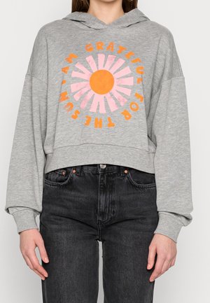 Osoba ubrana w szarą krótka bluzę z kapturem z różowo-pomarańczową grafiką słońca i tekstem "GRATEFUL FOR THE SUN I AM" pomarańczowymi literami.