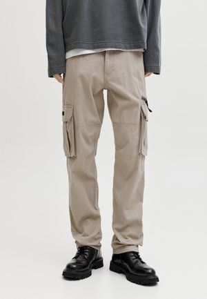 Άτομο που φοράει μπεζ παντελόνι cargo με πλαϊνές τσέπες, μαύρα παπούτσια με κορδόνια και σκουρόγριστο μακρυμάνικο πουκάμισο, στέκεται μπροστά σε απλό φόντο.