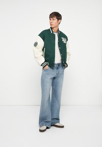 Grøn og creme varsityjakke med et rundt mærke, stribede manchetter og en tætsiddende silhuet, parret med lyseblå vide jeans og sneakers.
