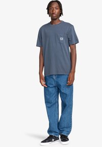 Mörkgrå kortärmad t-shirt med bröstficka, tillsammans med löst sittande blå denimjeans och svarta sneakers med vita sulor.