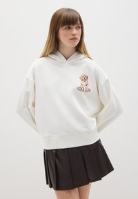 Witte hoodie met een grafische print aan de voorkant van een hond en de tekst "COOL CLUB", gecombineerd met een zwarte plooienleren rok. Zacht materiaal, relaxte pasvorm.