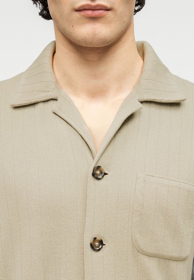 Μπεζ πουκάμισο με κουμπιά, με ριμπ υφή, γιακά με σχισμή και μικρή τσέπη στο στήθος, με στρογγυλά καφέ κουμπιά.