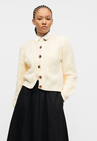 Νεαρή γυναίκα με κοτσίδες, που φοράει κρεμ μάλλινο ζακέτα με κουμπιά πάνω από λευκό πουκάμισο με γιακά και μαύρη φούστα, μπροστά από απλό λευκό φόντο.
