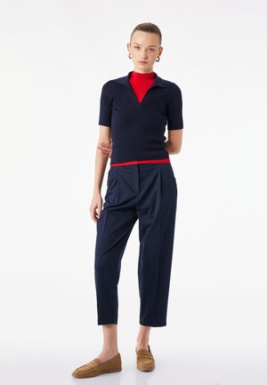 Kvinde står med armene bag ryggen iført marineblå kortærmet top over rød højhalset trøje, marineblå korte bukser og tanfarvede loafers.