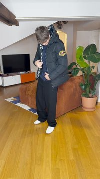 Schwarze Daunenjacke mit glänzender Oberfläche, ausgestattet mit einem Logopatch am linken Arm und einem kontrastierenden Innenmuster. Getragen über einem marineblauen Hemd.