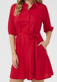 Czerwona sukienka koszulowa z zapięciem na guziki z przodu, krótkimi elastycznymi rękawami i paskiem wiązanym w talii. Charakteryzuje się rozkloszowanym dołem i bocznymi kieszeniami.