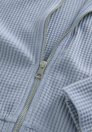 Lichtblauwe zip-up jas met een gestructureerd wafelpatter, voorzien van een zilverkleurige rits en zachte stof. Onafgewerkte manchetten en zoom.