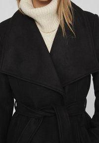 Cappotto a portafoglio nero con ampio colletto e vita incinta da cintura, realizzato in tessuto morbido e strutturato, indossato sopra un maglione a collo alto color crema.