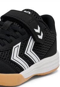 Chaussure de sport noire avec des bandes en chevron blanches, dessus en mesh, strap en velcro, lacets noirs, semelle intermédiaire blanche et semelle extérieure en caoutchouc beige.