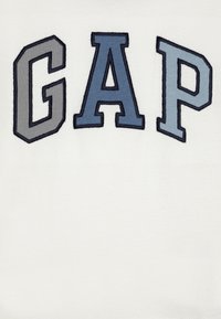 White fabric with the embroidered letters "GAP" in varying shades of blue and gray. The letters have a raised texture and clear outlines.