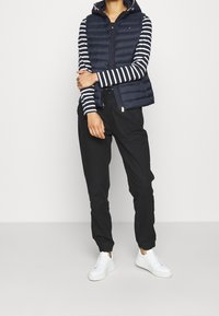 Henkilö, jolla on päällään laivastonsininen vuorattu huppupusakka musta-valkoisen raidallisen pitkähiiressä paidan, mustat jogger-housut ja valkoiset lenkkarit, seisoo.