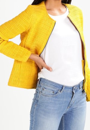 Kvinde iført en tekstureret gul jakke over en hvid t-shirt og lyseblå jeans med den ene hånd hvilende på hoften.