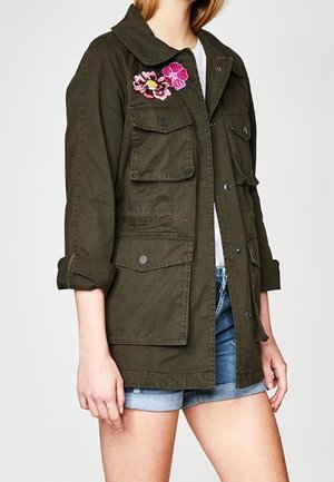 Donna che indossa una giacca verde scuro con quattro tasche frontali e ricami floreali rosa sul petto, abbinata a pantaloncini di jeans.