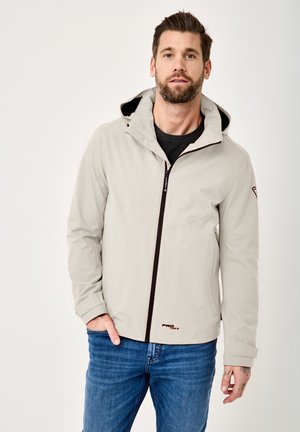 Mann trägt hellgraue Kapuzenjacke mit schwarzem Reißverschluss und blaue Jeans, steht mit einer Hand in der Tasche vor einfarbigem Hintergrund.