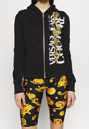 Vrouw die een zwarte hoodie draagt met witte "Versace Jeans Couture" tekst en een gouden barok design, gecombineerd met zwart-goudkleurige shorts met patroon.