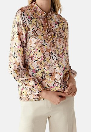 Kvinde iført en langærmet blomsterbluse med pink, brun, gul og sort farver, kombineret med lyse beige bukser, hænder samlet foran.