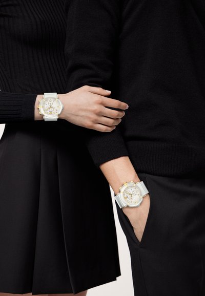 Dos relojes exhibidos: uno con una correa blanca y detalles en dorado, y otro con una correa gris y detalles dorados similares, usados sobre ropa oscura.