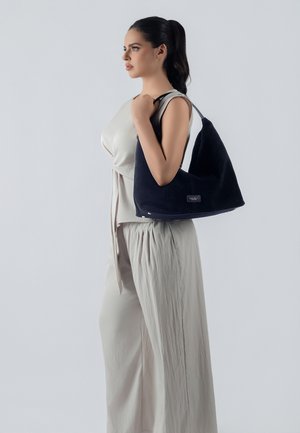 Hobo bag in navy suede with a single shoulder strap, featuring a smooth texture and subtle logo detail. Model wears light beige clothing.