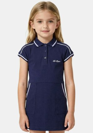 Jeune fille aux cheveux blonds portant une robe polo bleu marine avec des bordures blanches et des poches avant, posant devant un fond uni.