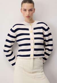 Pamučna prugasta kardigan u krem i mornarskoj plavoj boji, s okruglim izrezom, dugim rukavima i zlatnim dugmadima sprijeda. Jednostavna bež suknja.