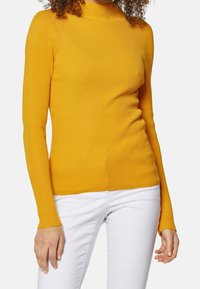 Personne portant un pull à col roulé côtelé jaune à manches longues, associé à un pantalon blanc, debout devant un fond uni.