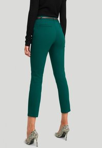 Zelené kalhoty na míru se slim fit střihem, středně vysokým pasem a bočními ventily, doplněné vysokými hadími podpatky a černým topem.