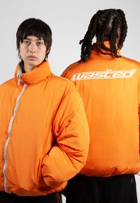 Chaqueta naranja acolchada con alto cuello y cremallera frontal. Presenta un audaz logo "wasted" en la parte posterior. Textura suave, diseño oversized.