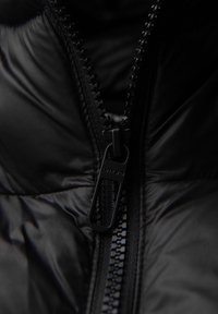 Černá péřová bunda vyrobená z látky s výrazným zipem. Zip má matný povrch a táhlo je označeno jako "ombre."