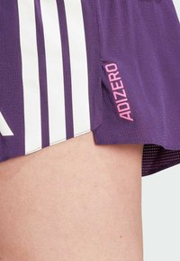 Ljubičaste atletske kratke hlače s bijelim horizontalnim prugama, s "ADIZERO" logotipom u ružičastoj boji sa strane i teksturiranim, laganim materijalom.