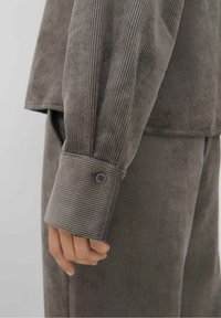 Camicia in velluto a coste grigia con una vestibilità strutturata, caratterizzata da costine verticali strette, polsini con bottone e una texture liscia. Messa in risalto da una leggera lucentezza.