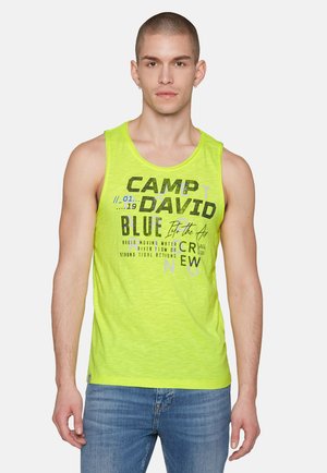 Helle gelbe Tanktop mit strukturierter Oberfläche, versehen mit schwarz und blau bedruckter Schrift. Kombiniert mit hellblauen Jeans.