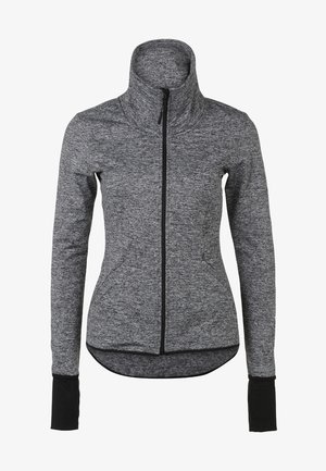 Osoba ubrana w szarą kurtkę na zamek, czarne spodnie z kieszenią na zamek oraz brązowe buty turystyczne, stojąca na jednolitym jasnym tle.