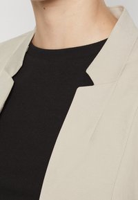 Beżowa marynarka z ostrym kołnierzem na czarnym, prążkowanym topie. Gładka tekstura tkaniny i dopasowany krój z czystymi liniami.