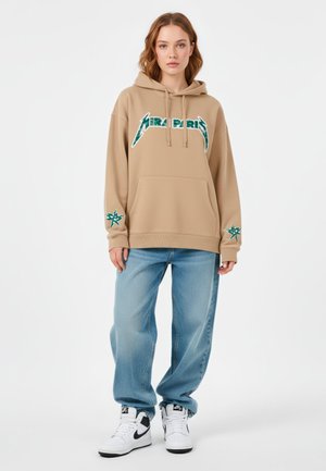 Felpa oversized beige con logo verde e accenti a stella sulle maniche, abbinata a jeans baggy blu chiaro e sneaker bianche.