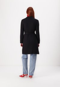 Manteau noir long avec une ceinture, présentant un col. Porté avec un jean bleu clair et des escarpins rouges. Texturé lisse, design simple.