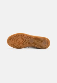 Semelle d'une chaussure en caoutchouc marron avec des motifs d'adhérence circulaires et ondulés sur un fond blanc uni.