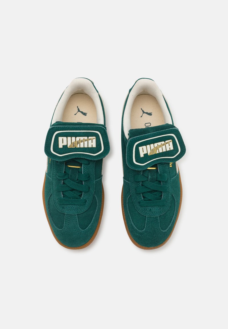 Sneaker in suede verde con striscia in velcro e lacci, caratterizzate da un interno bianco, suola in gomma e un evidente marchio "PUMA" in oro e bianco.