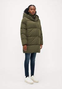 Doudoune vert olive avec un col haut et une capuche, présentant un design matelassé. Portée sur un jean bleu foncé et des baskets blanches à plateforme.