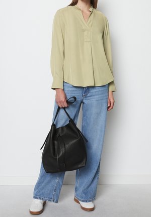 Personne portant un chemisier vert clair et un jean large bleu, tenant un grand sac seau en cuir noir par sa bandoulière.