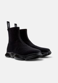 Botas negras hasta el tobillo con un upper de tejido elástico y una suela de goma lisa con bultos redondeados, equipadas con lengüetas para facilitar su colocación.