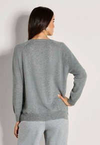 Frau mit langen dunklen Haaren, die einen grauen Strickpullover und graue Hose trägt, steht mit dem Rücken zugewandt und hat eine Hand auf der Hüfte.