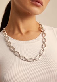 Collana in argento con maglie allungate e intrecciate, indossata su una maglietta bianca a coste. Texture liscia e finitura lucida.