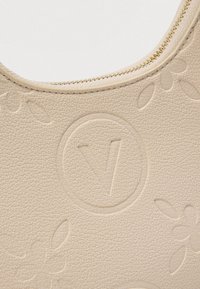 Bež teksturirana usnjena površina z reliefnimi cvetličnimi vzorci in krogom, v katerem je stilizirana črka "V" blizu zlatega zadrge.