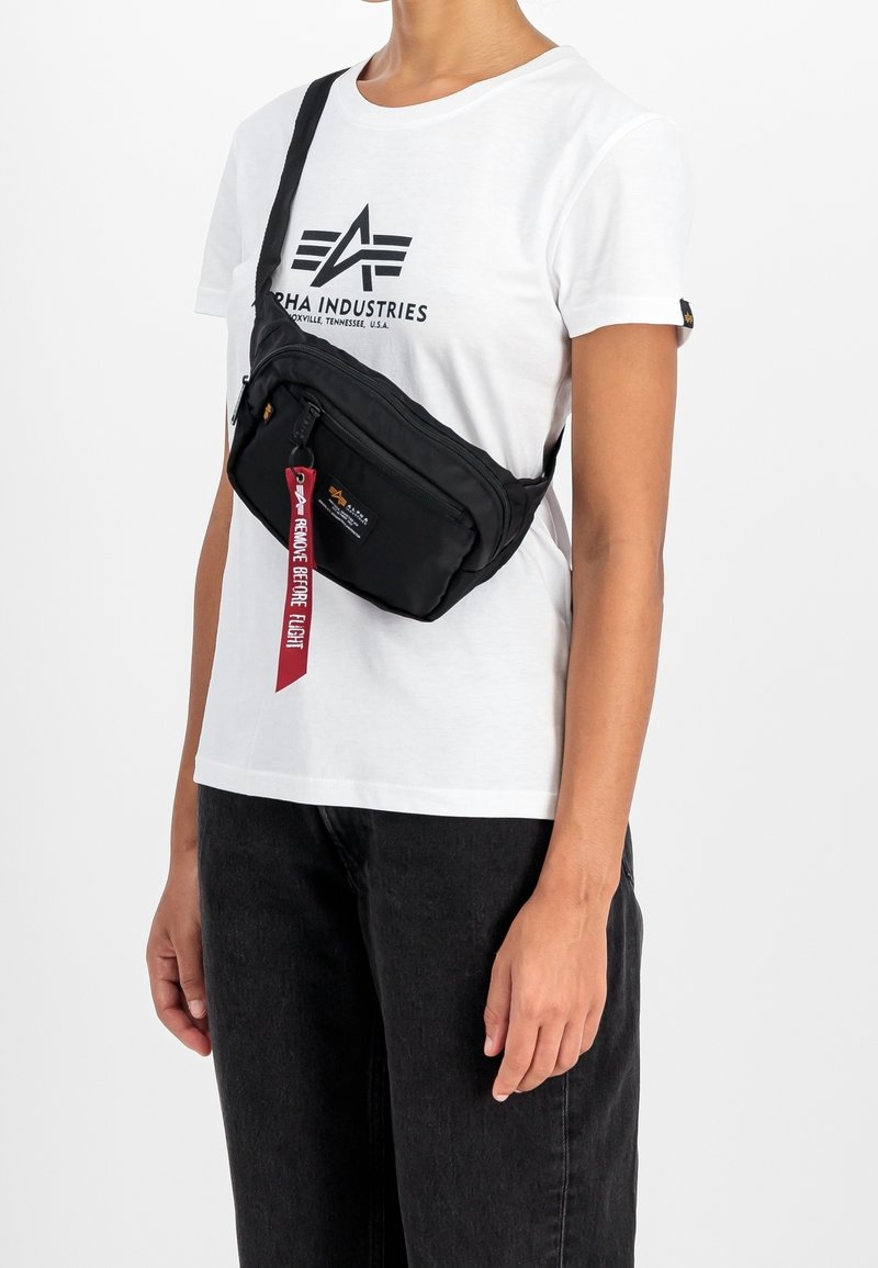 Personne portant un t-shirt blanc Alpha Industries et un sac bandoulière noir avec une étiquette rouge indiquant "Remove Before Flight,", associé à un pantalon noir.