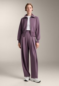 Lila Zip-Jacke mit kurzem Kragen, kombiniert mit hoch taillierten, weit geschnittenen Hosen. Glattes Material mit lässiger Passform. Weißes T-Shirt darunter.