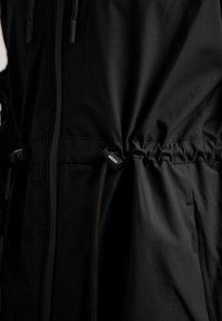 Giacca esterna nera con tessuto liscio, zip frontale, cordoncino regolabile in vita e dettaglio con logo.